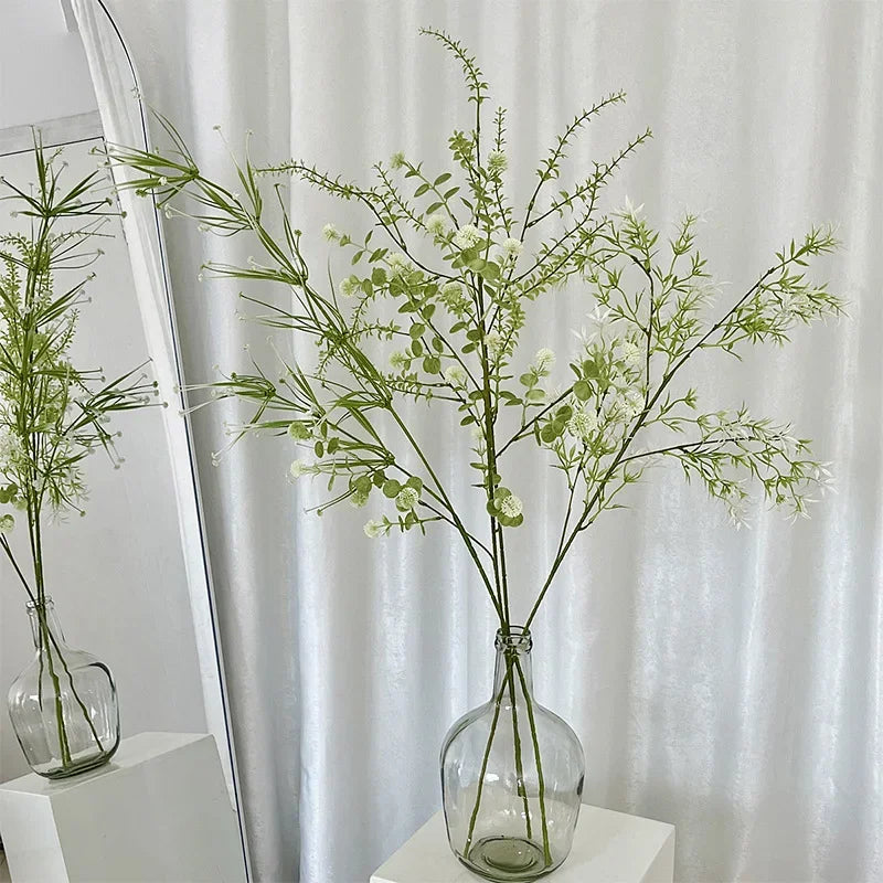 Clear glass vase with green branches against a white curtain background