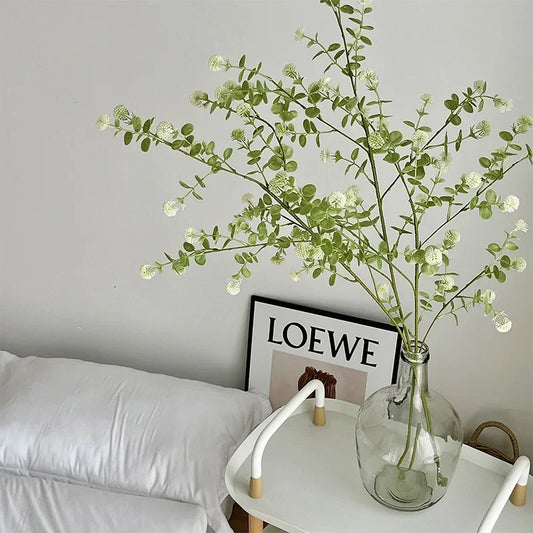 Decorative green branches in a clear vase on a side table with a LOEWE poster in the background.
