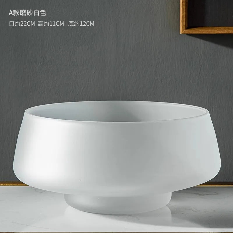 A frosted glass vase in a bronze color, displayed on a shelf with a small bouquet of flowers inside it, alongside a round glass bowl and a framed picture in the background.